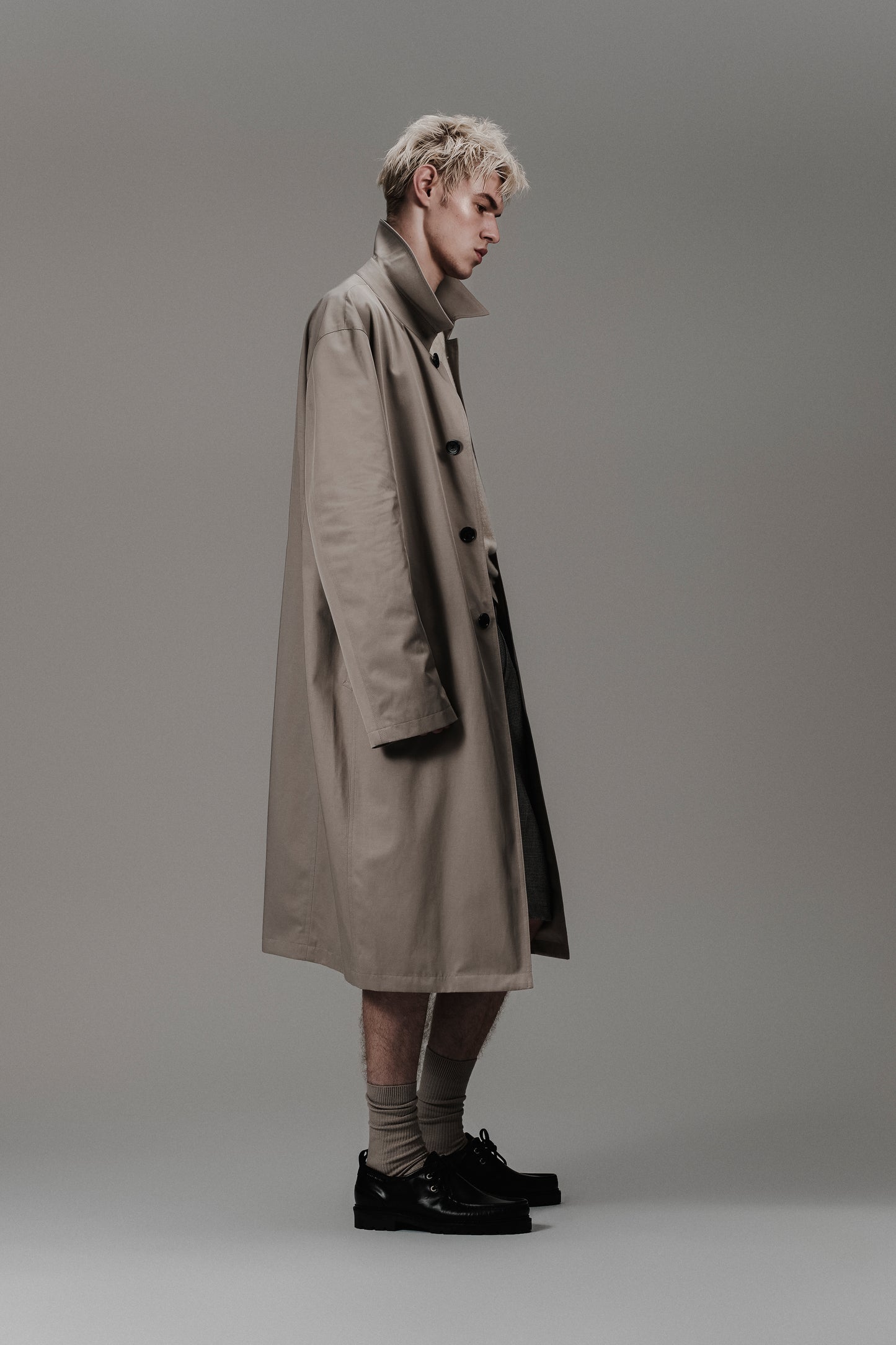 Side profile of a Celine model wearing oversized trench coat in technical fabric, savile row style, white vest and wool hounstooth long shorts. Inspired by Balenciaga, vtmnt and margiela.