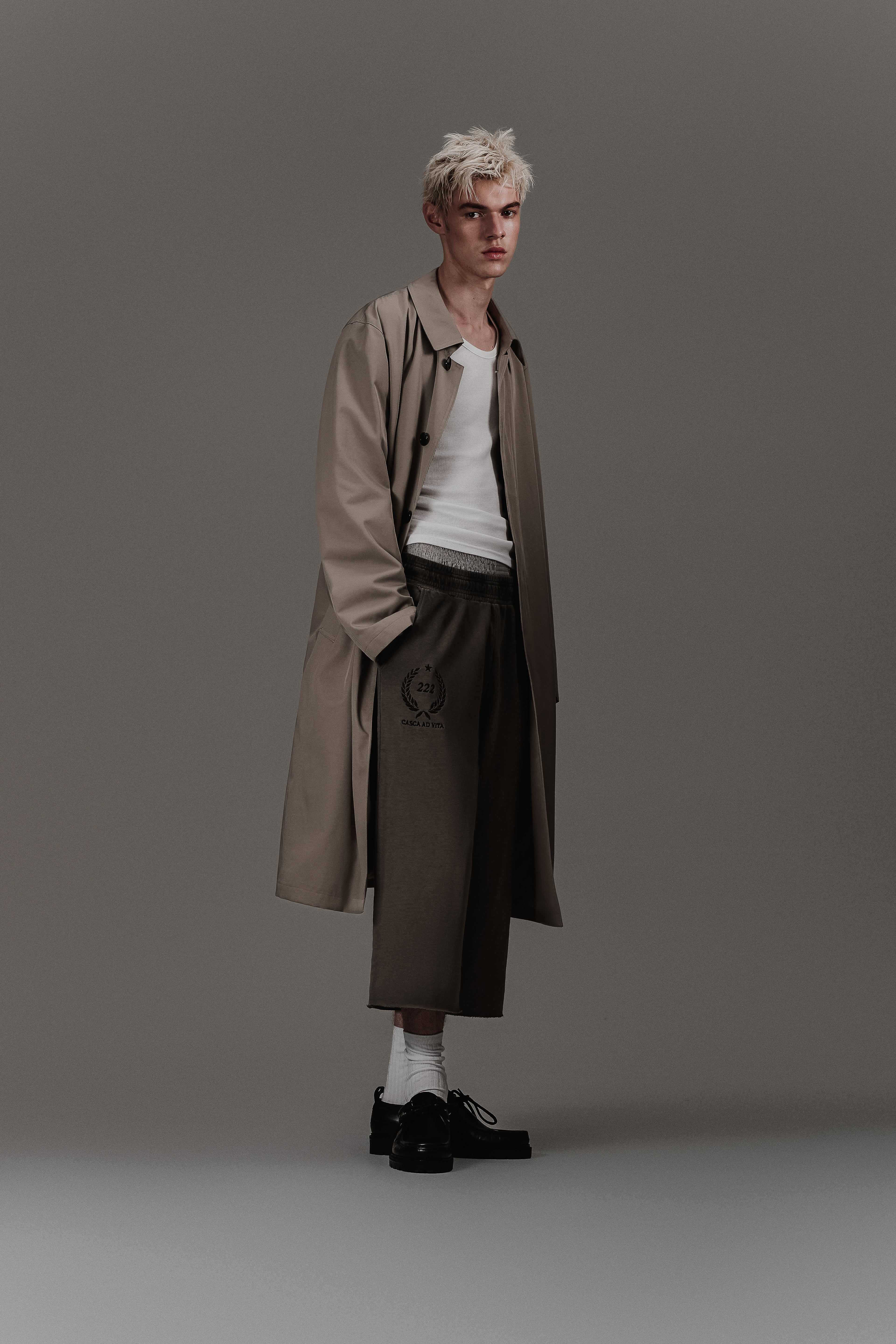 Celine model wearing a pair of oversized faded grey sweatpants with an elastic waistband, embroidered 222 crest logo and embroidered casca 222 sport logo. Raw cut hem for a cropped fit. Oversized trench coat, white vest. Inspired by Balenciaga, vtmnt and margiela.