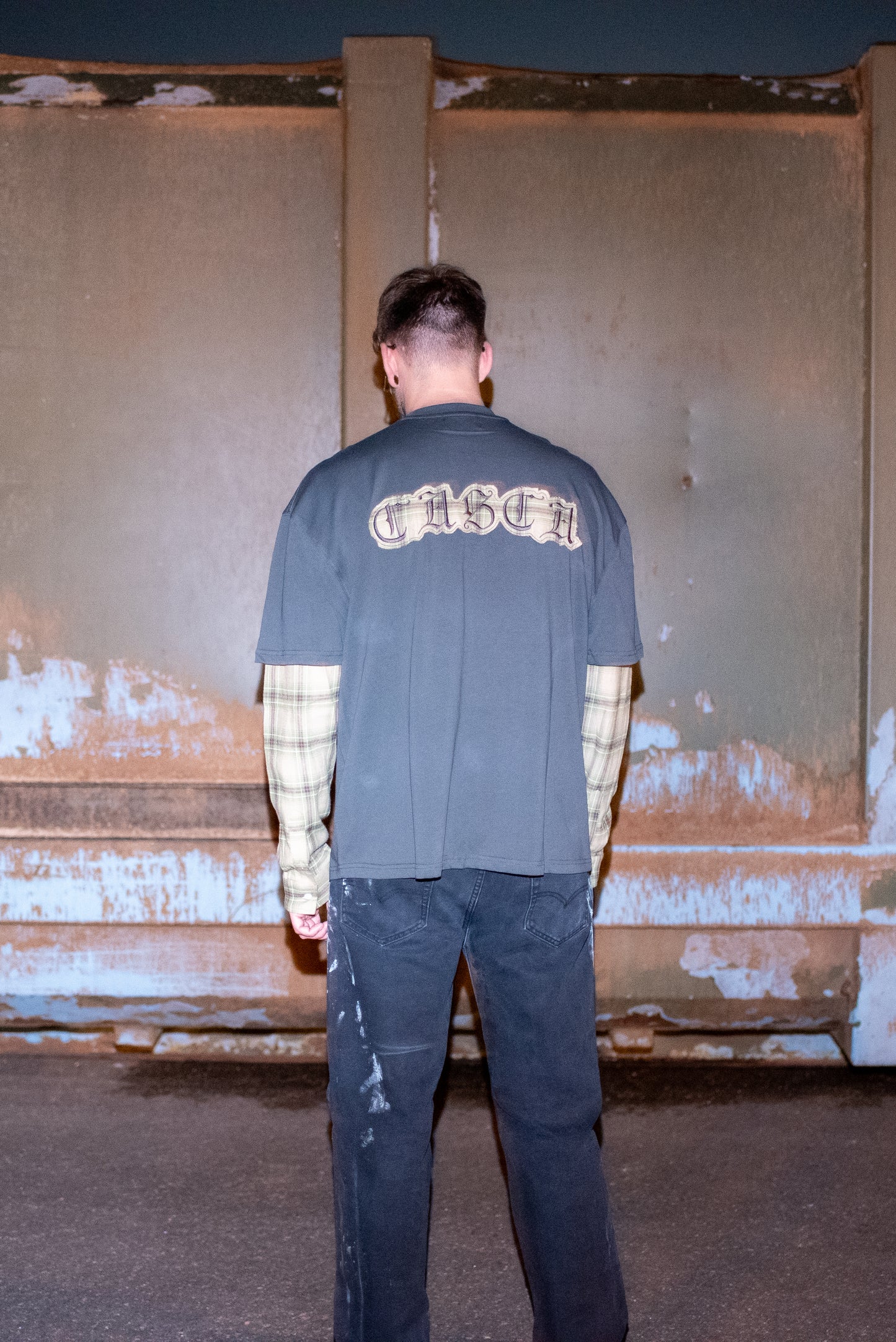 Oversized T-shirt in grey with plaid sleeves worn by model in Chrome Hearts Glasses, customised painted levi's jeans and Baleniaga shoes, shown from the back