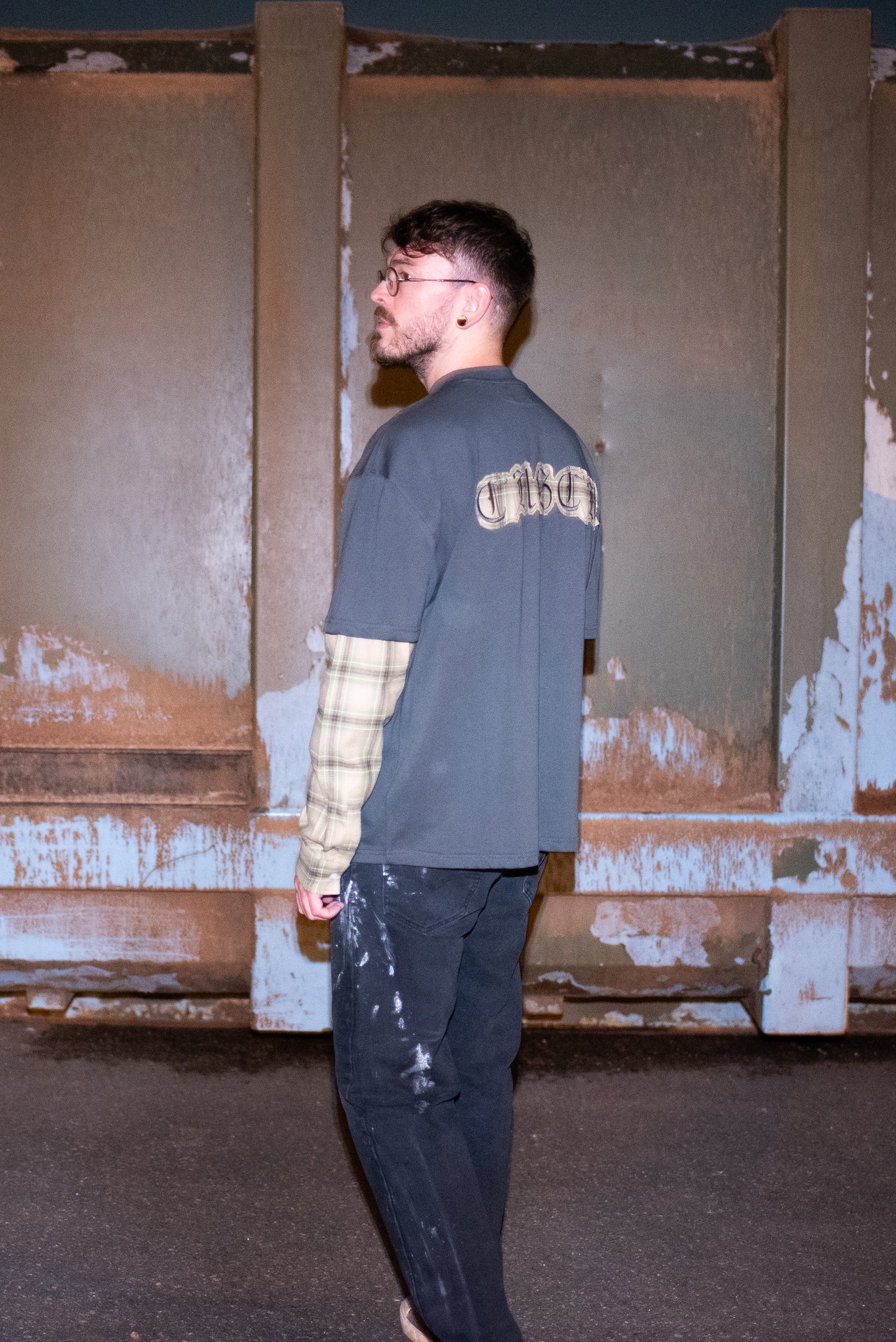 Oversized T-shirt in grey with plaid sleeves worn by model in Chrome Hearts Glasses, customised painted levi's jeans and Baleniaga shoes, shown from the side