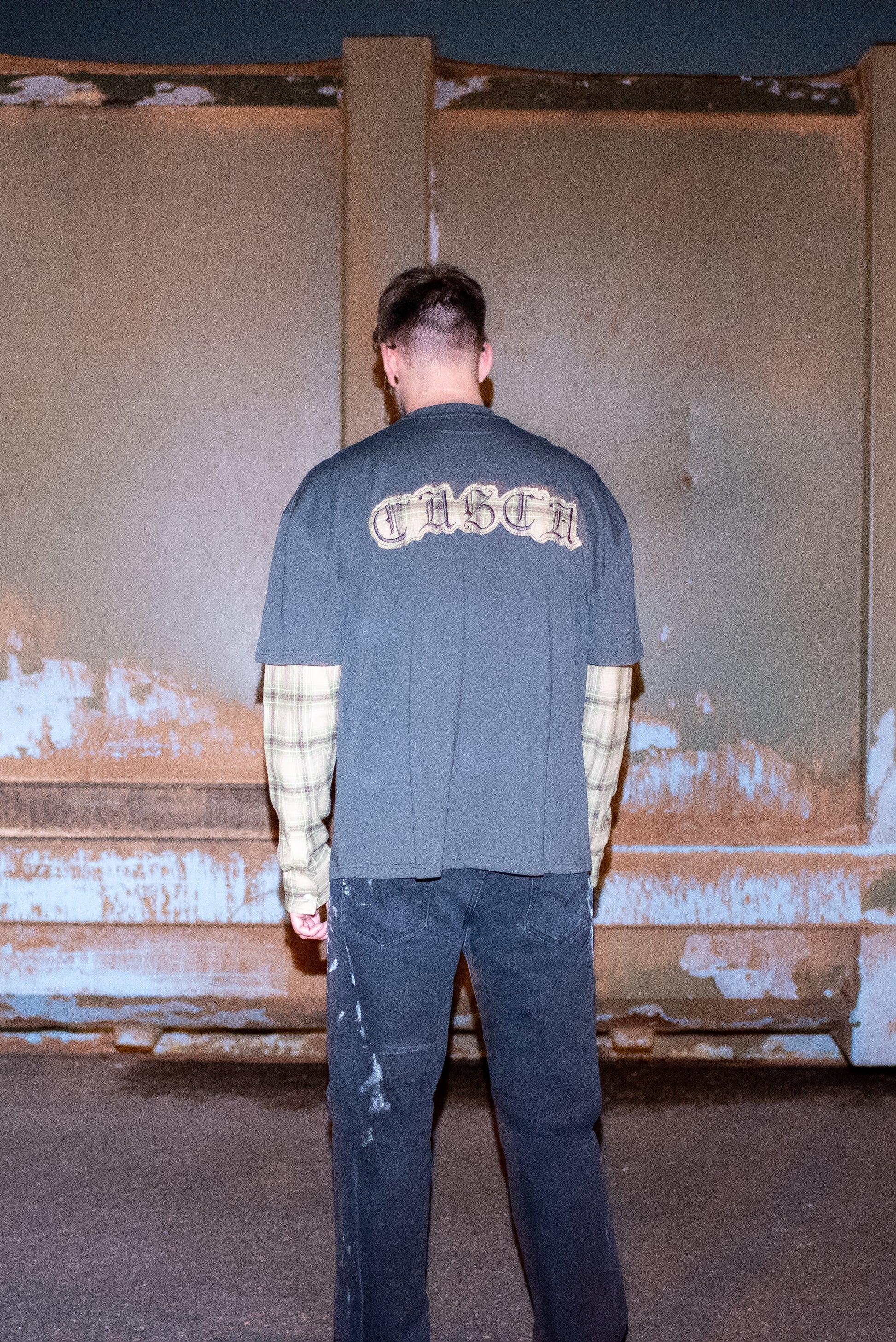 Oversized T-shirt in grey with plaid sleeves worn by model in Chrome Hearts Glasses, customised painted levi's jeans and Baleniaga shoes, shown from the back