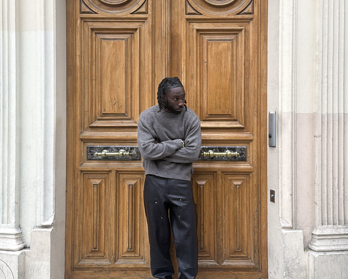 kojey radical standing in front of a large wooden door with classical architecture. wearing leather double knee trouser and wool sweater.